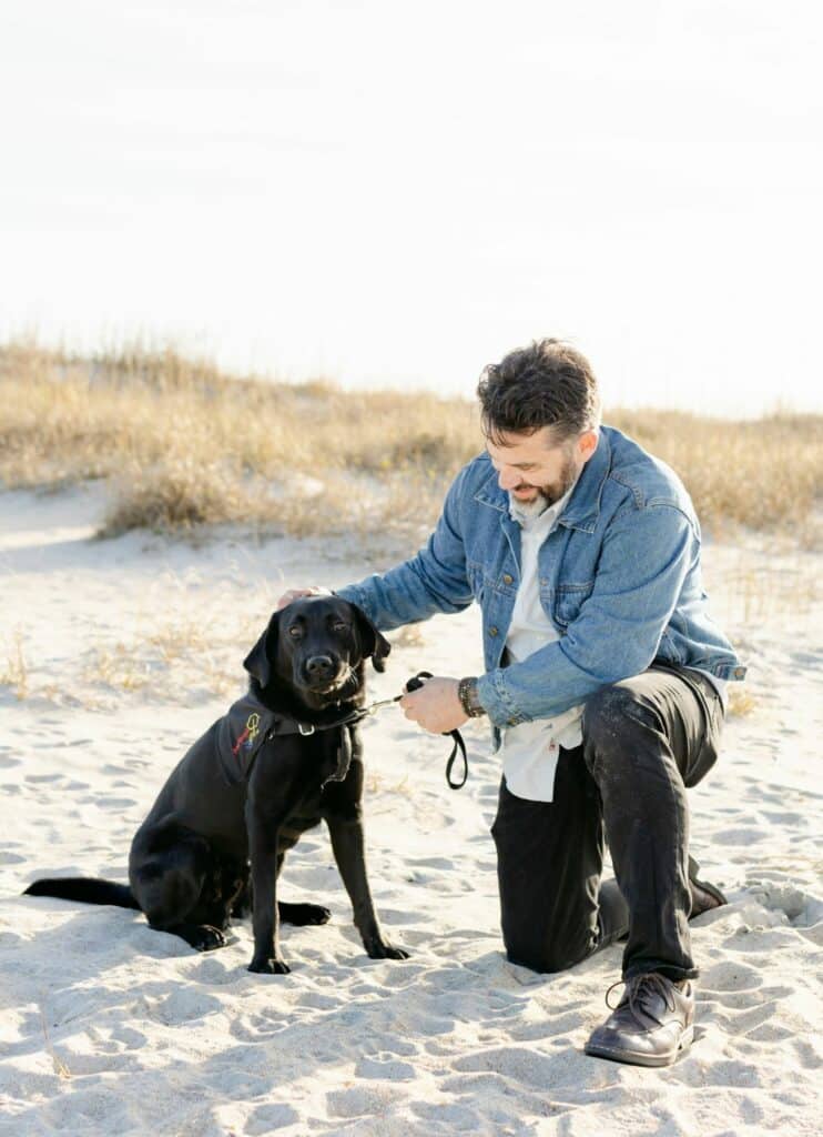 black lab service dog in vest sitting on beach with handler kneeling next to it